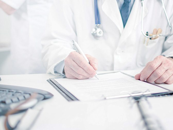 A clinician in a white coat with a stethoscope writing on a patient chart